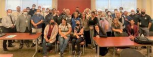 A large group photo of a diverse team of people gathered in a conference room, with some wearing masks.