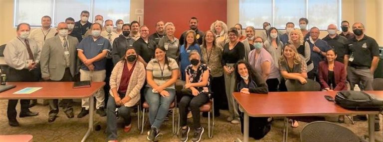A large group photo of a diverse team of people gathered in a conference room, with some wearing masks.