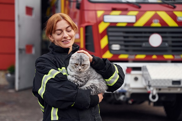 A female firefighter in uniform holding a gray Scottish Fold cat, standing in front of a fire truck.