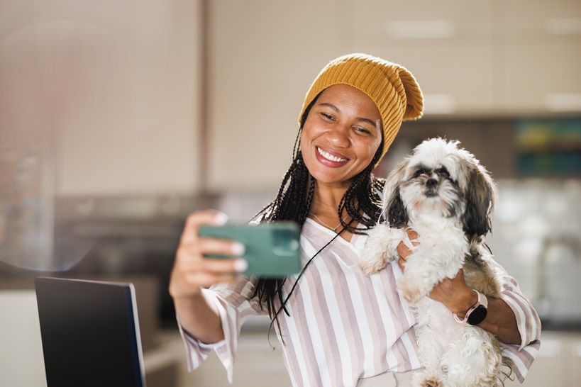 A smiling woman holding a small fluffy dog while taking a selfie in a bright indoor setting.