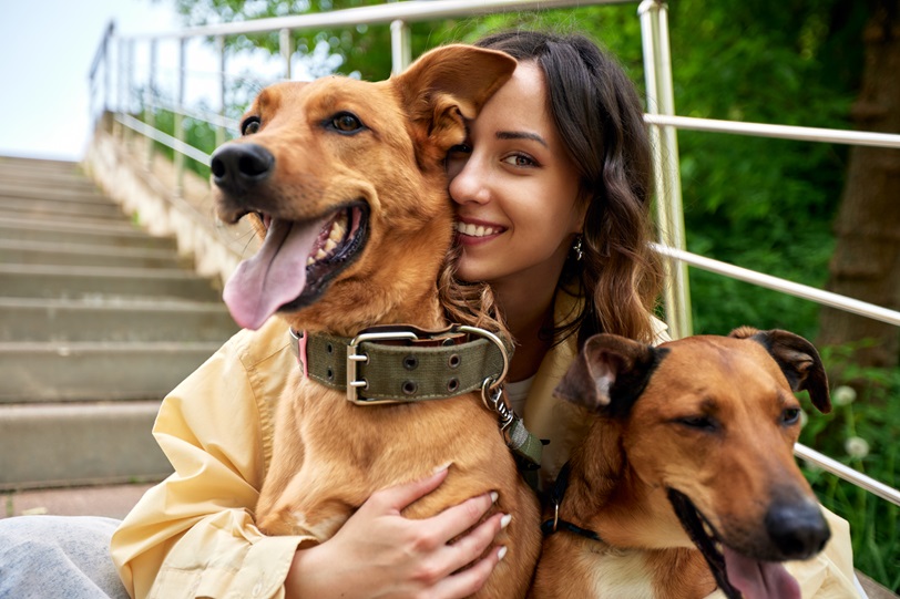 A smiling woman sitting on outdoor steps with two happy brown dogs. She is hugging the dogs, and all appear joyful and relaxed, with greenery in the background.