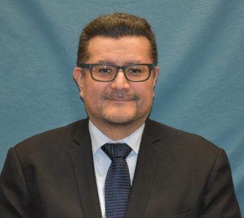 "Portrait of Samuel Ortiz, Departmental Information Security Officer, wearing a dark suit, blue patterned tie, and glasses, standing against a blue backdrop