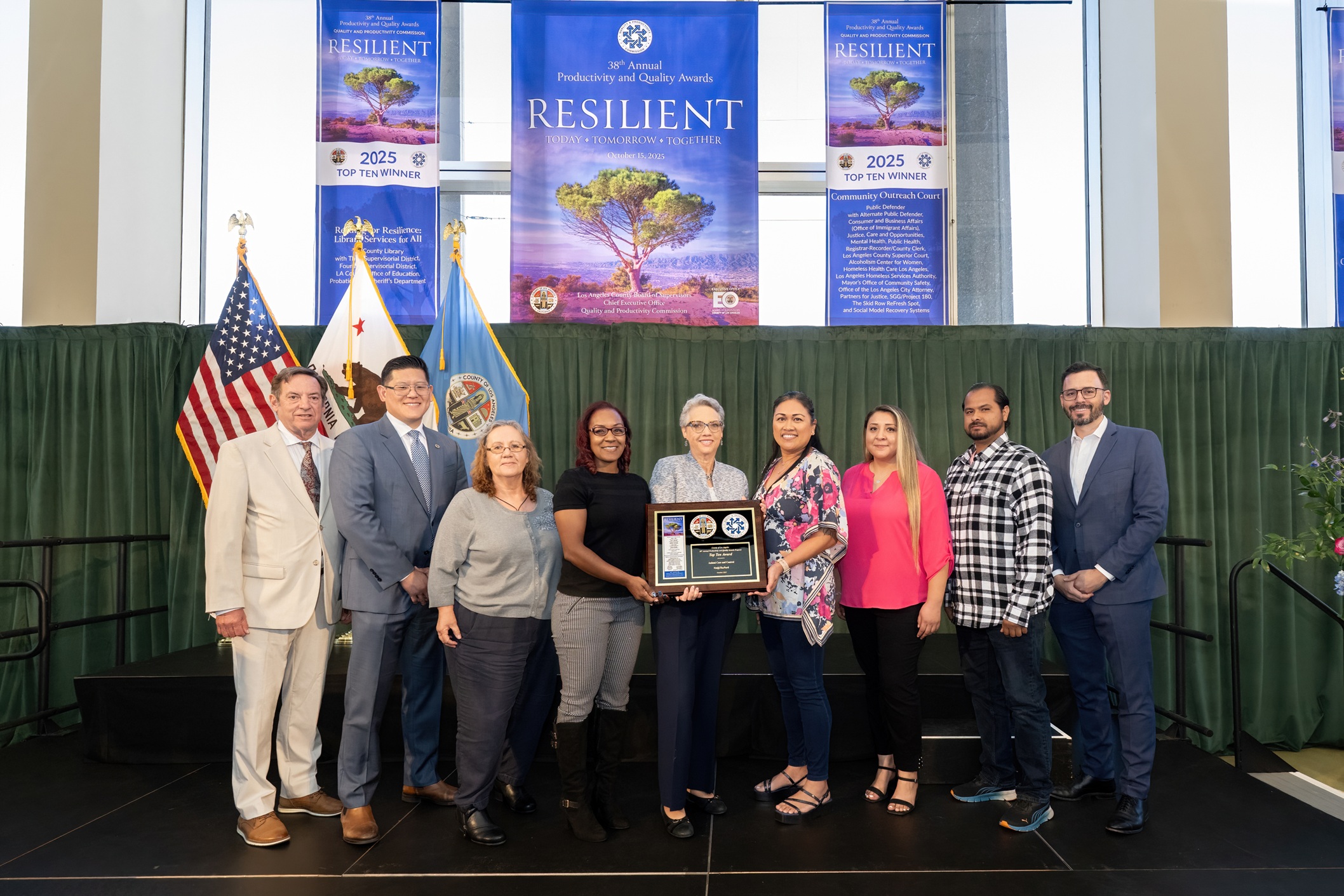 A group of eight people stand on a stage holding a framed award plaque. They are posing for a photo in front of banners that read “2025 Top Ten Winner” and “Resilient — Today, Tomorrow, Together.” The American flag, the California state flag, and the Los Angeles County flag are displayed behind them. Everyone is smiling, dressed in professional or business-casual attire, and standing in front of a green curtain backdrop.