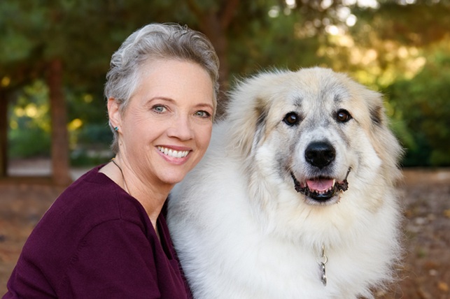 Smiling woman with short gray hair posing outdoors beside a large, fluffy light-colored dog, both facing the camera with trees softly blurred in the background